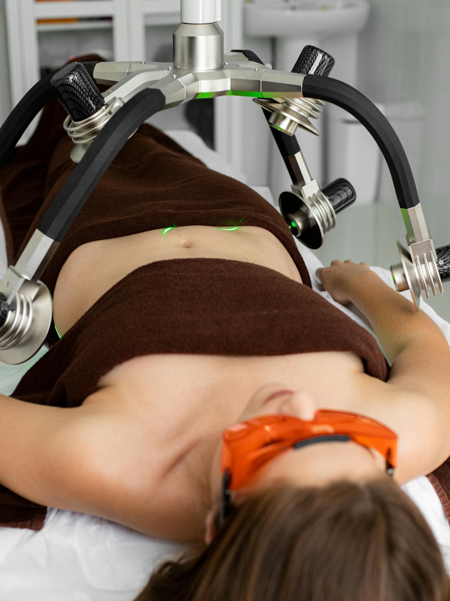 Female patient covered with towels in protective eyeshades lying under modern laser weight loss machine during treatment in medical clinic
